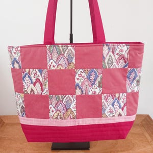 May include: A pink quilted tote bag with a patchwork design. The bag features a mix of floral and paisley patterns. The handles and base are a darker pink, while the main body has a lighter pink background. The bag is suitable for shopping.