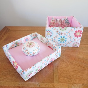 May include: A white fabric box with a pink floral pattern and a pink fabric lid. The lid has a small pink and white fabric cushion in the center.