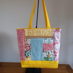 May include: A yellow patchwork tote bag with a zipped pocket. The bag is made of different fabrics with floral and leaf patterns in shades of red, green, blue, and white.
