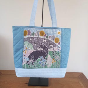 May include: A blue quilted tote bag featuring a fox and cows in a field print. The print includes a fox, cows, trees, and flowers in shades of green, blue, yellow, and pink.