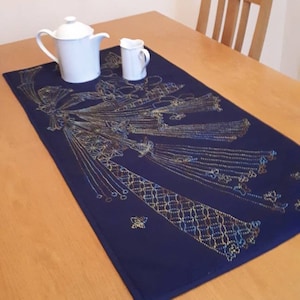 May include: A navy blue table runner with a gold, yellow, and blue embroidered design. The runner is on a wooden table with a white teapot and creamer.