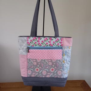 May include: A patchwork tote bag with a grey base and pink, blue, and white floral patterns. The bag has a zipped pocket and two grey handles.