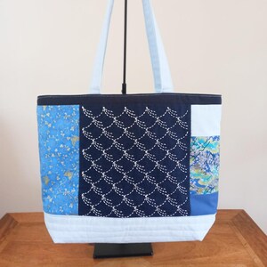 May include: A blue and white tote bag with a patchwork design. The bag has a large central panel with a navy blue background and white stitching in a wave pattern. The other panels feature a blue floral pattern, a blue and gold floral pattern, and a blue and green floral pattern.