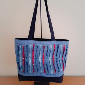 May include: A blue tote bag with a decorative front panel featuring vertical strips of blue and red fabric. The bag has navy blue handles and a navy blue base. The bag is displayed on a black stand.