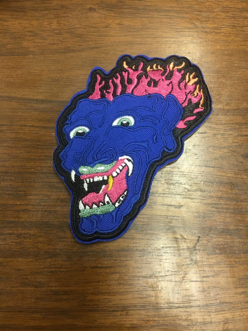 Pu&ograve; includere: Toppa ricamata raffigurante un volto di mostro blu con occhi verdi, bocca rosa e denti bianchi. Il mostro ha fiamme rosa e arancioni sulla parte superiore, delineate con cuciture nere. La toppa &egrave; su una superficie di legno.