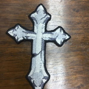 May include: Embroidered cross patch in shades of gray and white with a black border. The cross has a detailed, textured design, and is likely for clothing or accessories. The patch is on a wooden surface.