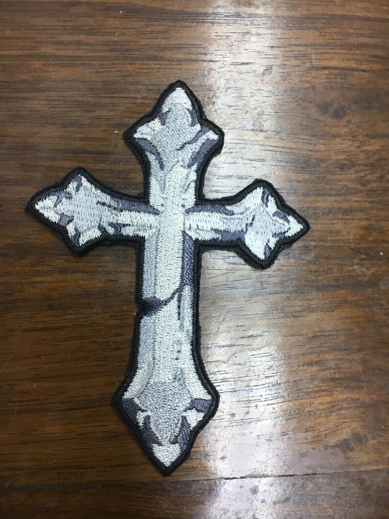 May include: Embroidered cross patch in shades of gray and white with a black border. The cross has a detailed, textured design, and is likely for clothing or accessories. The patch is on a wooden surface.