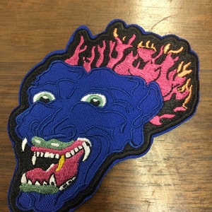 Pu&ograve; includere: Toppa ricamata raffigurante una faccia di mostro blu con occhi verdi, denti bianchi e una lingua rosa. La testa del mostro ha fiamme rosa e arancioni. La toppa &egrave; bordata di nero.