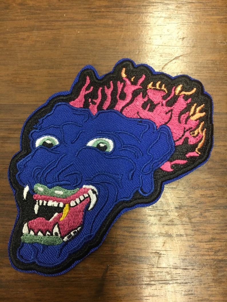 Pu&ograve; includere: Toppa ricamata raffigurante una faccia di mostro blu con occhi verdi, denti bianchi e una lingua rosa. La testa del mostro ha fiamme rosa e arancioni. La toppa &egrave; bordata di nero.