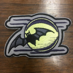 May include: Embroidered patch featuring a black bat silhouetted against a yellow moon, set within a dark purple and gray border. The design includes stylized clouds and a textured appearance, suitable for jackets or bags.
