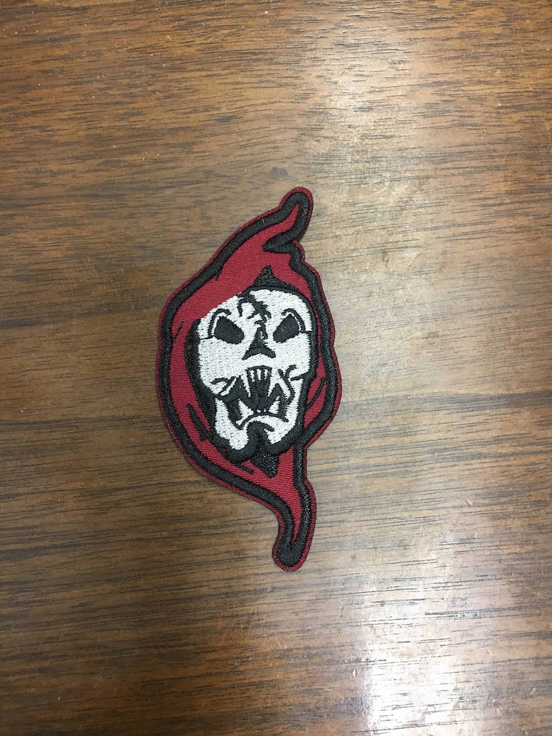 May include: Embroidered patch featuring a white skull with sharp teeth, set against a red hooded cloak design. The patch has a black border and is likely for clothing or accessories. The skull has black details.