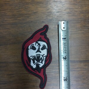 May include: Embroidered patch depicting a skull with a red hood and black edging. The patch is approximately 7.6 cm in height. The skull has white and black details, including sharp teeth. The patch is on a wooden surface.