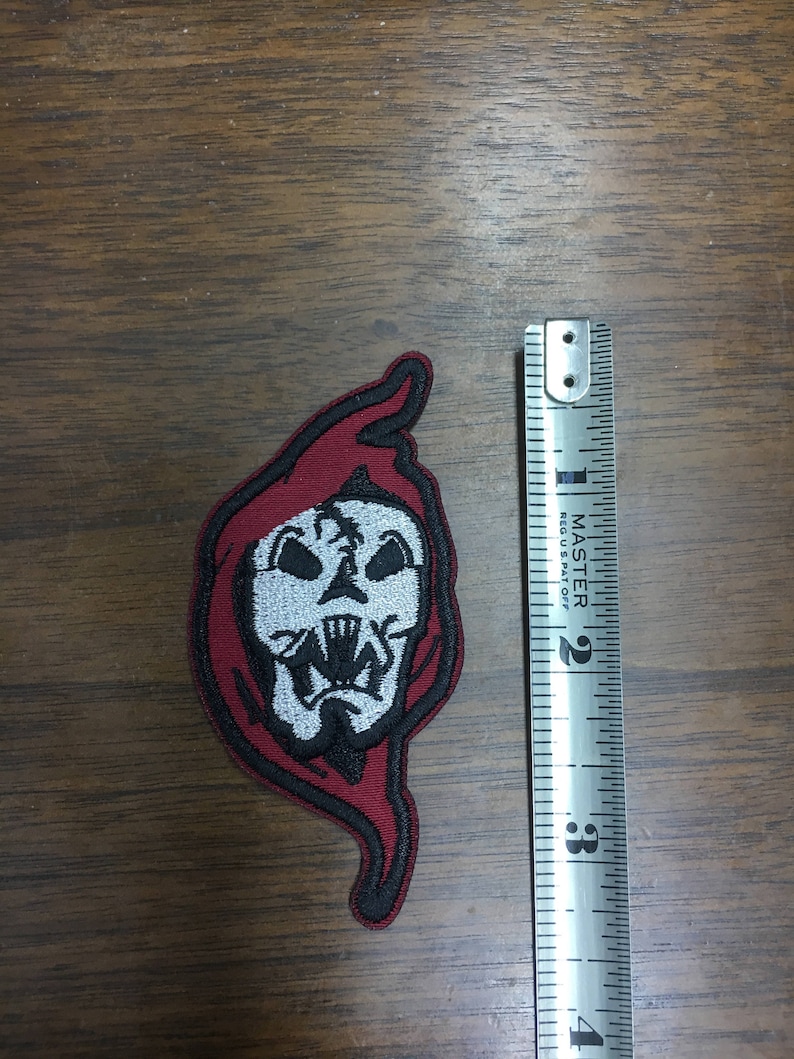 May include: Embroidered patch depicting a skull with a red hood and black edging. The patch is approximately 7.6 cm in height. The skull has white and black details, including sharp teeth. The patch is on a wooden surface.