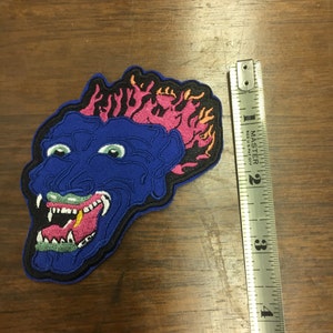 Pu&ograve; includere: Toppa ricamata raffigurante un volto di mostro blu con occhi verdi, bocca spalancata e fiamme rosa e gialle. La toppa &egrave; alta circa 10 cm. La toppa ha un bordo nero e si trova su una superficie di legno.