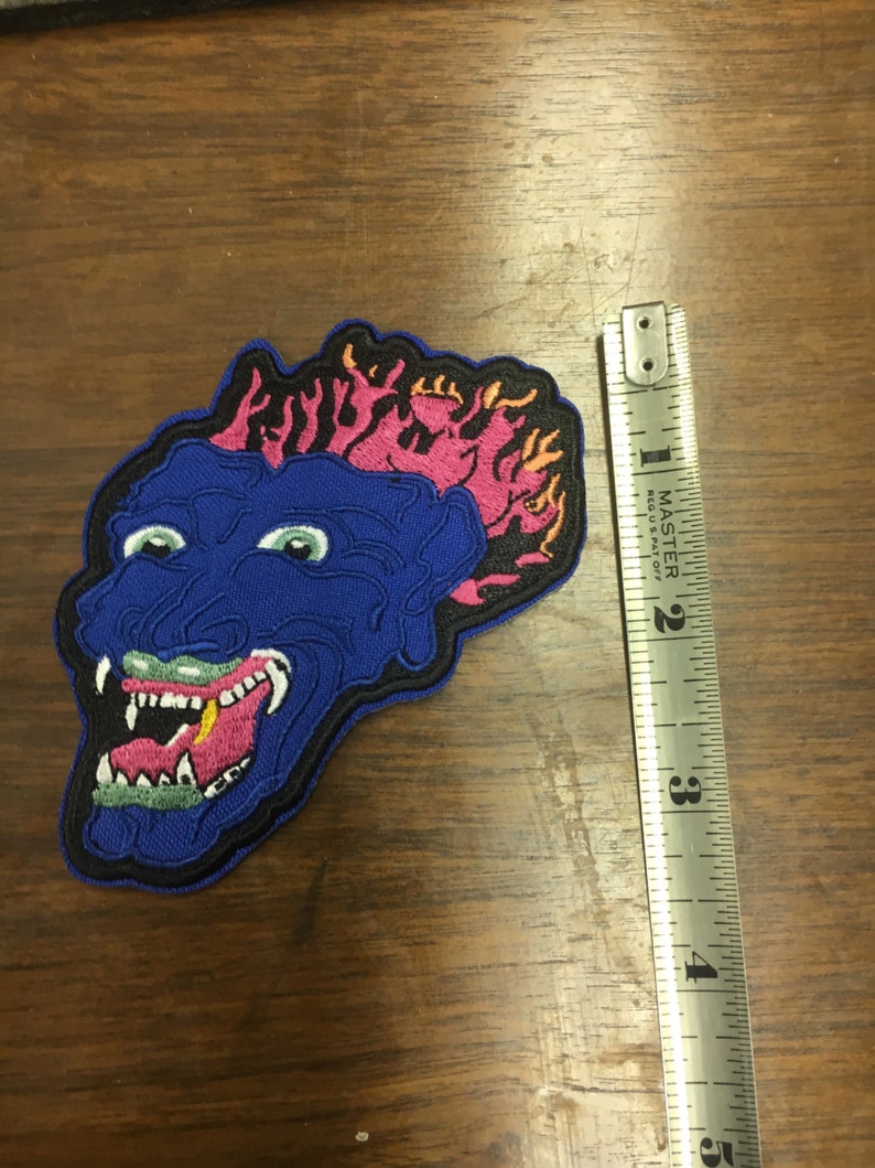 Pu&ograve; includere: Toppa ricamata raffigurante un volto di mostro blu con occhi verdi, bocca spalancata e fiamme rosa e gialle. La toppa &egrave; alta circa 10 cm. La toppa ha un bordo nero e si trova su una superficie di legno.