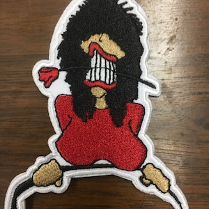 May include: Embroidered patch featuring a cartoon character with a large, toothy grin and wild black hair. The character wears a red top and has gold-colored hands. The patch has a white border and is likely designed for clothing or accessories.