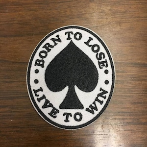 Pode incluir: Um patch redondo com um símbolo de espadas preto no centro. O patch branco tem texto bordado em preto que diz: "BORN TO LOSE. LIVE TO WIN."
