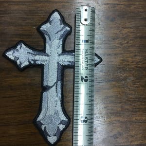 May include: Embroidered gray cross patch with a black border. The patch measures approximately 3 inches in height. The cross has a detailed, textured design, giving it a weathered appearance. The patch is on a wooden surface.