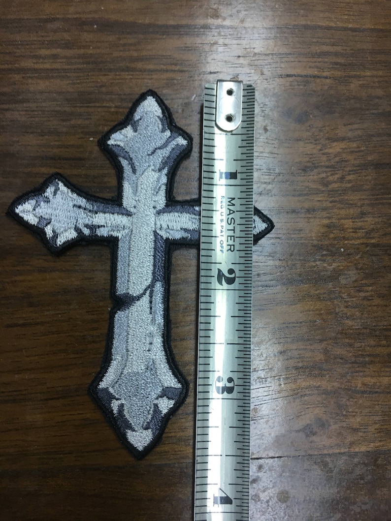 May include: Embroidered gray cross patch with a black border. The patch measures approximately 3 inches in height. The cross has a detailed, textured design, giving it a weathered appearance. The patch is on a wooden surface.