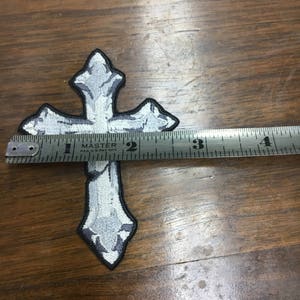 May include: Embroidered cross patch in shades of gray and white with a black border. The patch is approximately 3.5 inches long. The cross is measured with a metal ruler on a wooden surface. The patch has a decorative design.