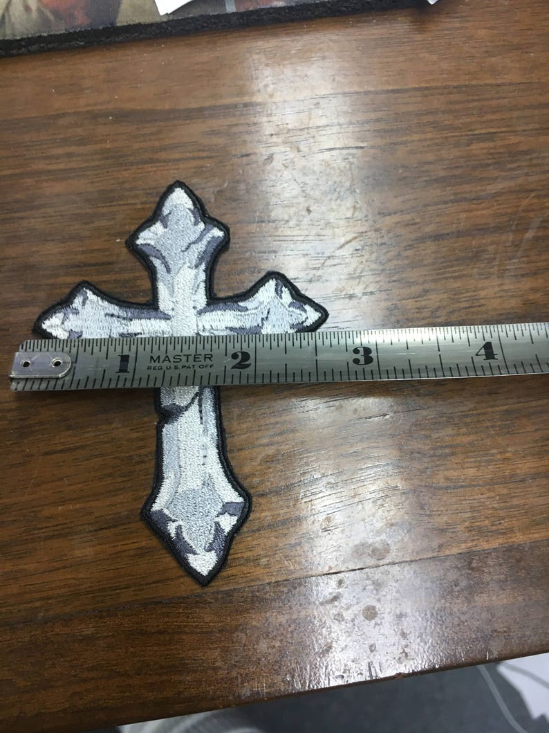 May include: Embroidered cross patch in shades of gray and white with a black border. The patch is approximately 3.5 inches long. The cross is measured with a metal ruler on a wooden surface. The patch has a decorative design.