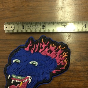 Pu&ograve; includere: Toppa ricamata con un volto di mostro blu con occhi verdi, una bocca spalancata e fiamme rosa. La toppa misura circa 7,6 cm di larghezza.