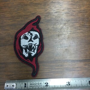 May include: Embroidered patch featuring a skull with a red hood and black outline. The skull is white with black details. The patch is approximately 5 cm in length.