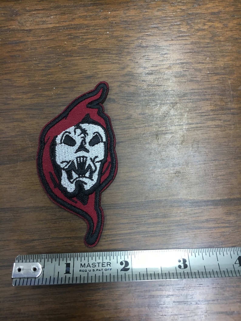 May include: Embroidered patch featuring a skull with a red hood and black outline. The skull is white with black details. The patch is approximately 5 cm in length.