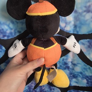 May include: A plush toy of a character with large black ears, wearing an orange pumpkin-shaped body suit with a brown stem on top, white gloves, and yellow shoes. The character has a black tail and a small tag attached to the lower back area.