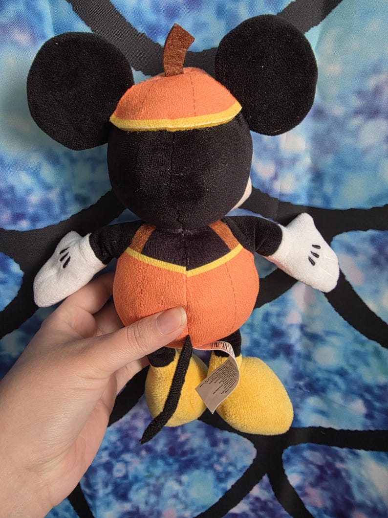 May include: A plush toy of a character with large black ears, wearing an orange pumpkin-shaped body suit with a brown stem on top, white gloves, and yellow shoes. The character has a black tail and a small tag attached to the lower back area.