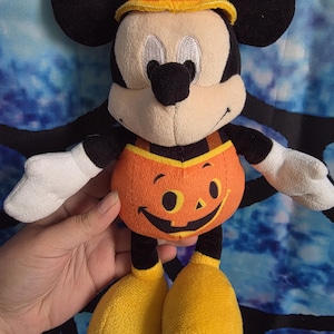 May include: A plush Mickey Mouse toy wearing a pumpkin-themed outfit. The character has black ears, a tan face, white gloves, and yellow shoes. The orange pumpkin costume features a printed jack-o'-lantern face and a matching orange pumpkin hat.