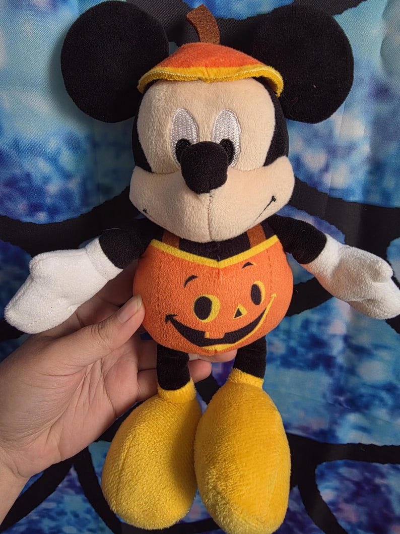 May include: A plush Mickey Mouse toy wearing a pumpkin-themed outfit. The character has black ears, a tan face, white gloves, and yellow shoes. The orange pumpkin costume features a printed jack-o'-lantern face and a matching orange pumpkin hat.