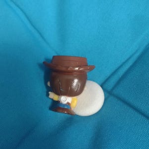 May include: A small, brown-haired figurine of a cowboy wearing a brown hat, yellow shirt, blue jeans, and white vest. The figurine is holding a white object. The background is a solid teal color.