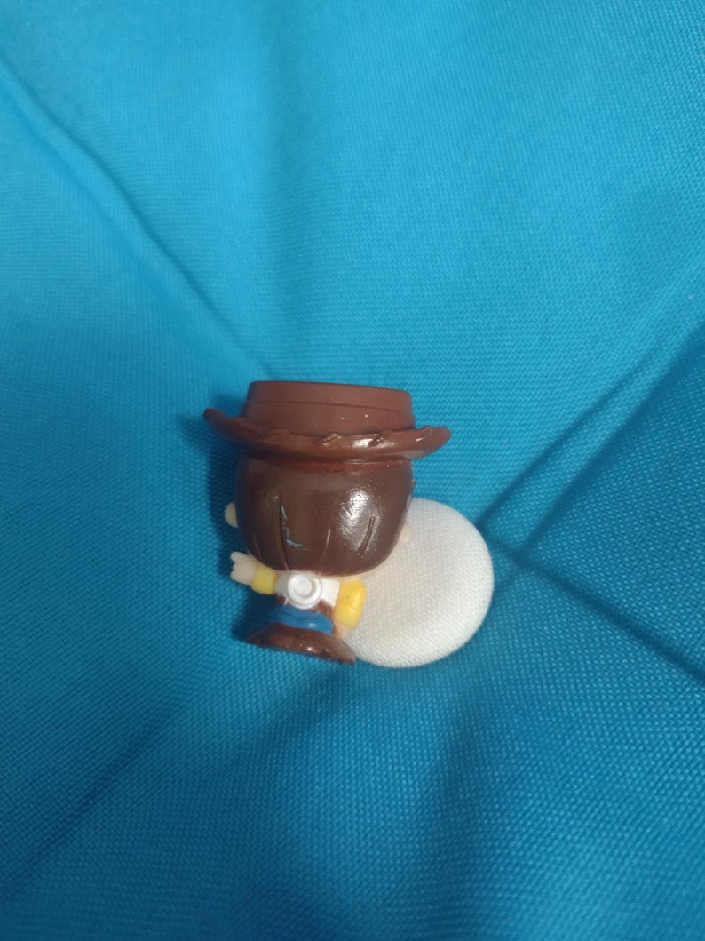 May include: A small, brown-haired figurine of a cowboy wearing a brown hat, yellow shirt, blue jeans, and white vest. The figurine is holding a white object. The background is a solid teal color.