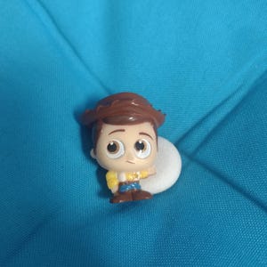 May include: A small, cartoon-style figure of Woody from Toy Story. The figure has a brown cowboy hat, brown hair, and a yellow shirt with a blue vest and pants. The figure is on a white circular base against a blue background.