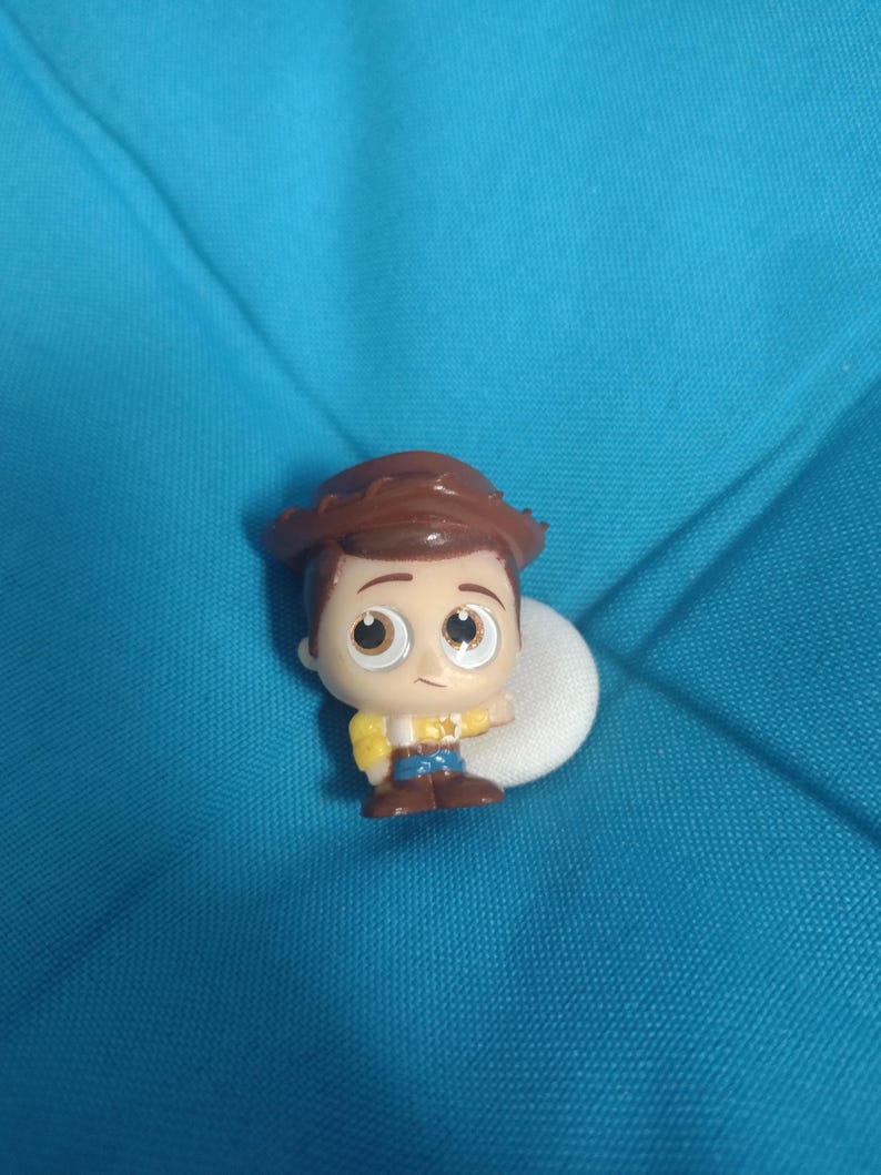 May include: A small, cartoon-style figure of Woody from Toy Story. The figure has a brown cowboy hat, brown hair, and a yellow shirt with a blue vest and pants. The figure is on a white circular base against a blue background.
