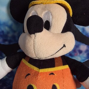 May include: A plush Mickey Mouse character toy wearing a pumpkin-themed outfit. The figure has black ears, a tan face, and a bright orange pumpkin body with a stitched jack-o'-lantern face. A yellow brimmed hat sits on top of the head.
