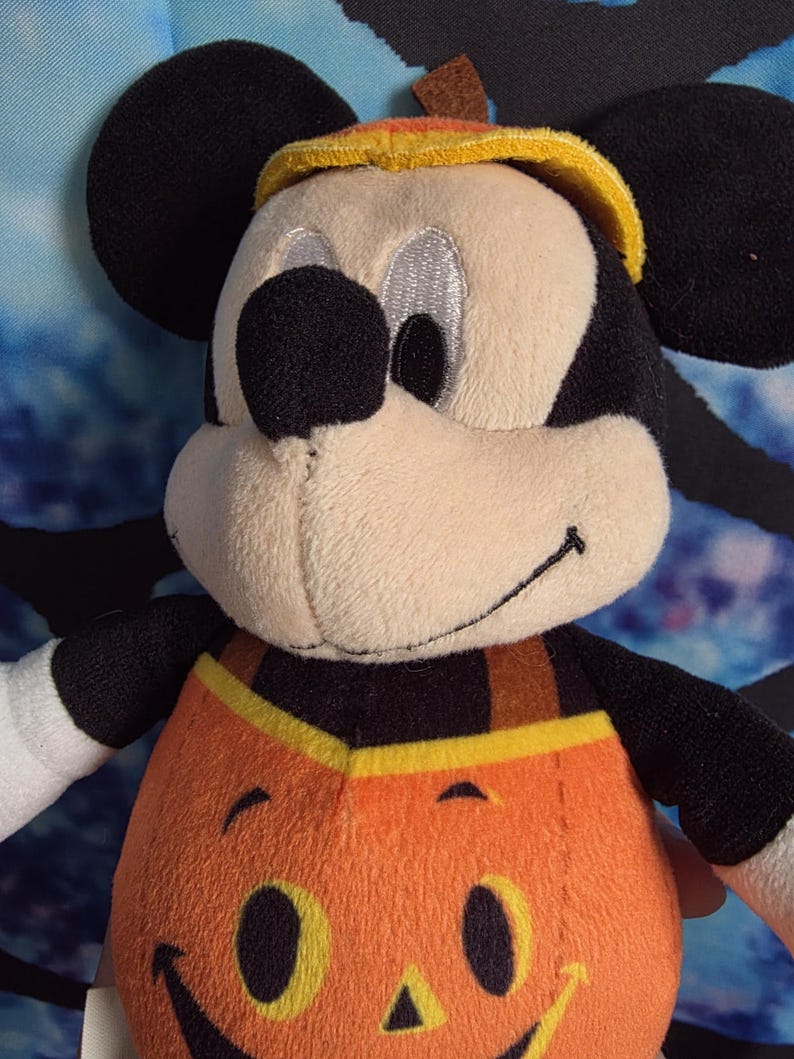 May include: A plush Mickey Mouse character toy wearing a pumpkin-themed outfit. The figure has black ears, a tan face, and a bright orange pumpkin body with a stitched jack-o'-lantern face. A yellow brimmed hat sits on top of the head.