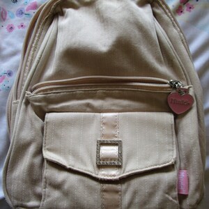 May include: A beige canvas backpack with a pink heart charm that says "Mud Pie". The backpack has a zippered pocket on the front with a silver buckle.