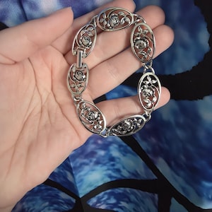 May include: Silver-toned bracelet featuring an oval link design with rose motifs. The bracelet is composed of multiple oval links, each adorned with a detailed rose and leaf pattern. The links are connected by smaller, rectangular connectors, creating a flexible and elegant piece of jewelry.