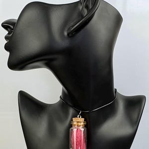 May include: A necklace featuring a small clear glass bottle pendant suspended from a thin black cord. The bottle is sealed with a cork stopper and contains a vibrant pink feather plume resting atop a layer of tiny, shimmering silver-toned beads.