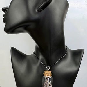 May include: A clear glass vial pendant filled with dried lavender flowers and small purple beads, sealed with a cork stopper. The pendant hangs from a simple black cord necklace, showcasing a botanical theme with natural textures and colors.