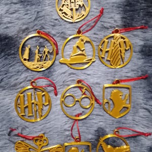 May include: Twelve gold-coloured Harry Potter themed Christmas decorations with red string hangers. The ornaments feature various Harry Potter symbols, including the Hogwarts crest, the Sorting Hat, the Deathly Hallows symbol, and the house crests.