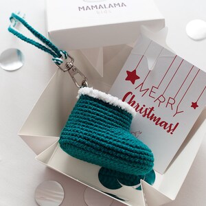 Peut inclure: Une botte en crochet bleu sarcelle avec une bordure en fourrure blanche, attachée à une ficelle blanche et bleu sarcelle avec un porte-clés argenté. La botte est dans une boîte blanche avec une carte qui dit "Joyeux Noël !"