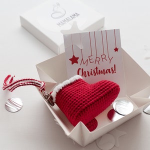 Puede incluir: Una pequeña media de Navidad roja de ganchillo con ribete blanco y una cinta a rayas blancas y rojas que cuelga de una caja de regalo blanca con una tarjeta que dice "¡Feliz Navidad!"