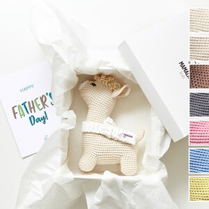 May include: A white gift box with a cream-colored crocheted llama toy inside. The box is filled with white tissue paper. A card with the text "Happy Father's Day!" is visible on the left side of the image. A small label with the text "MAMAL" is visible on the box.