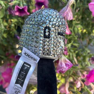 May include: A silver-colored, bedazzled Star Wars character ornament with a black face and a black handle. The ornament is covered in small, sparkling rhinestones. A white tag with text is attached to the handle.