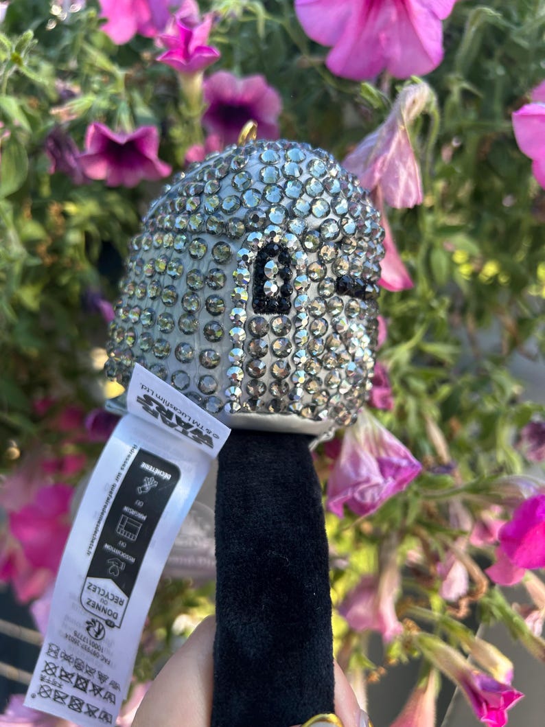 May include: A silver-colored, bedazzled Star Wars character ornament with a black face and a black handle. The ornament is covered in small, sparkling rhinestones. A white tag with text is attached to the handle.