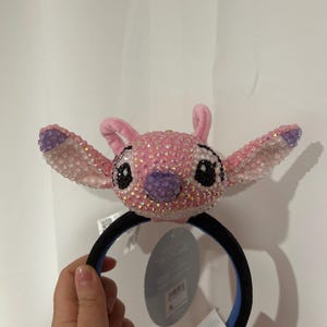 May include: A headband featuring a plush character head covered in pink and iridescent crystals. The character has large ears with purple accents, black crystal eyes, and a purple crystal nose. The headband band is black with a blue interior lining.