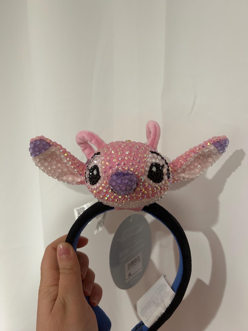 May include: A headband featuring a plush character head covered in pink and iridescent crystals. The character has large ears with purple accents, black crystal eyes, and a purple crystal nose. The headband band is black with a blue interior lining.